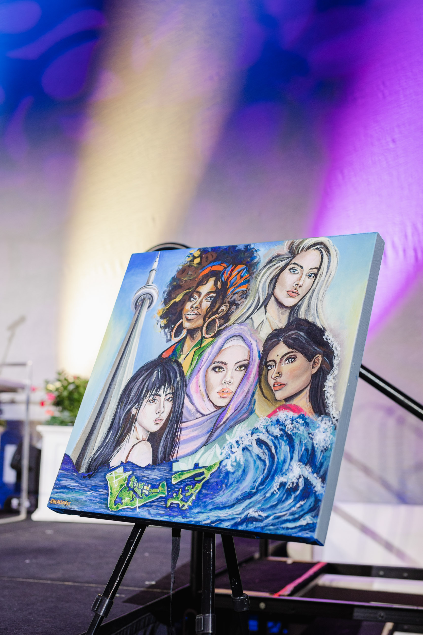 A conference photography of women posing in front of an easel with their painting displayed.