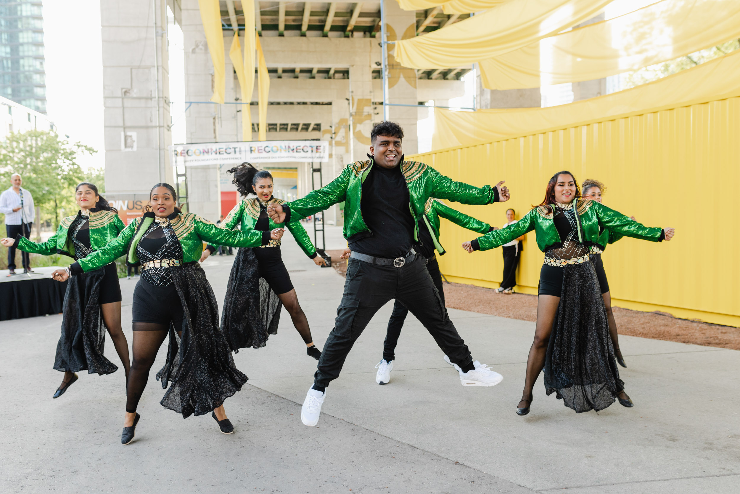 Conference Photography: A group of dancers in green jackets performing in front of a vibrant yellow building.