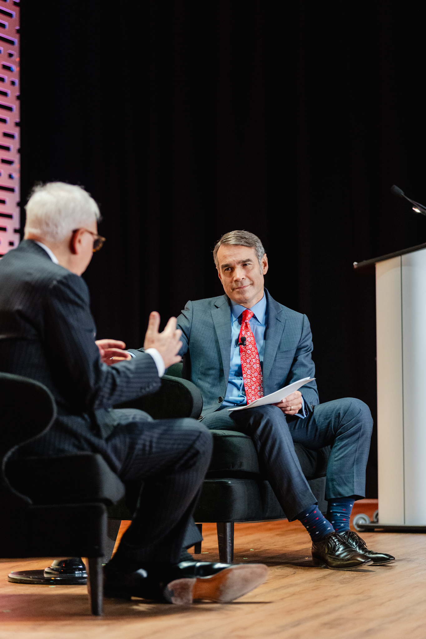 Conference photography of two men in suits engaging in conversation on stage.