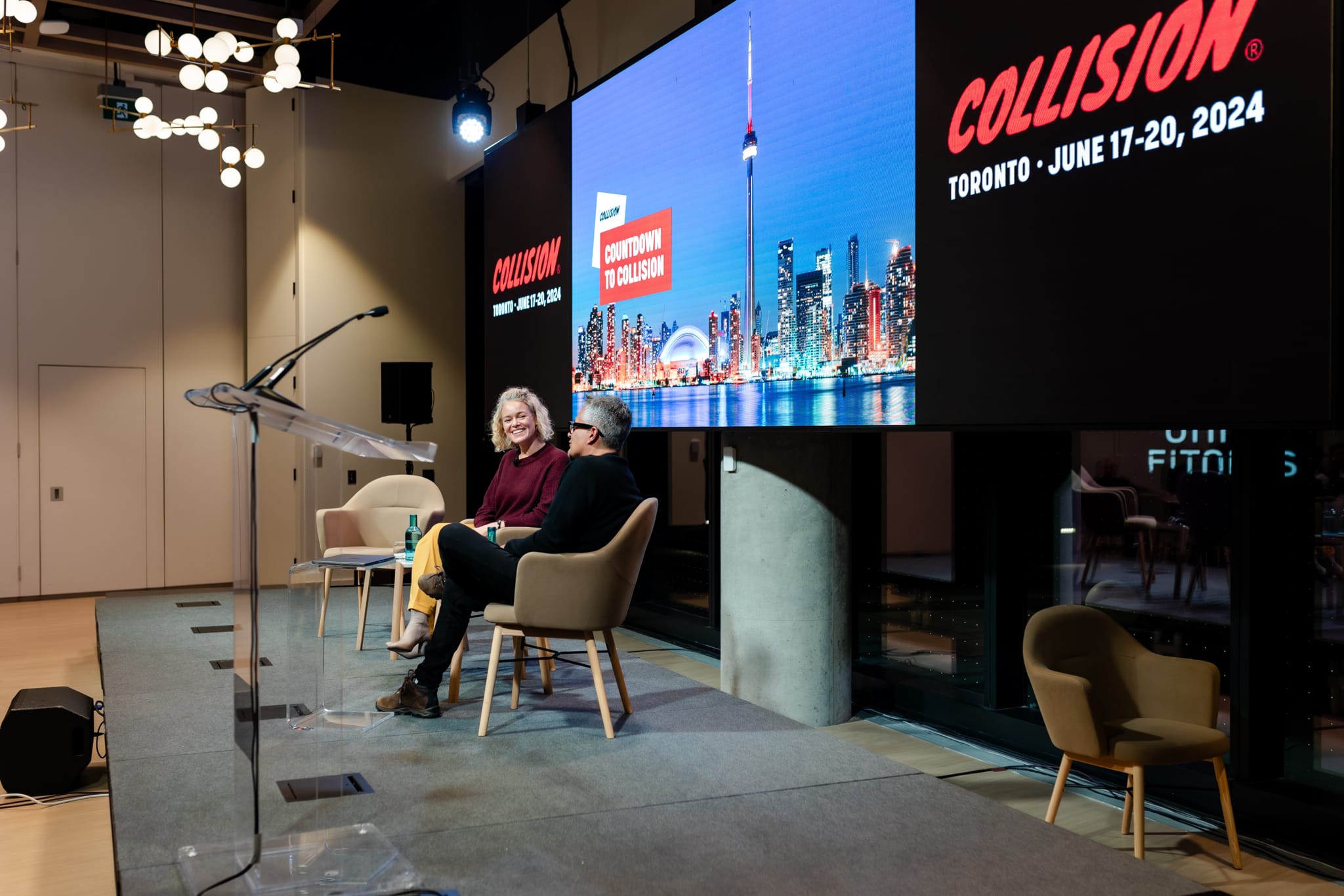 Two women seated on chairs in front of a large screen at an event.
