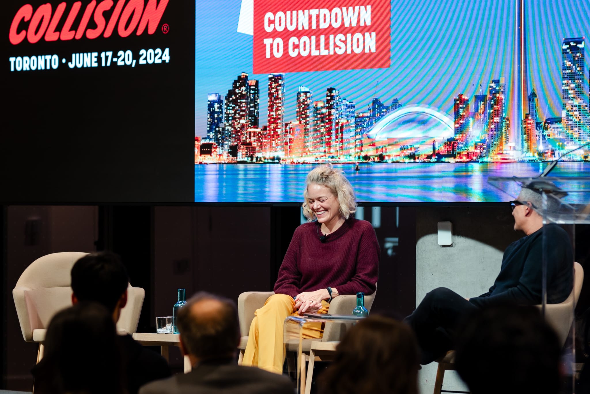 Two women are photographed sitting in front of a screen at an event.
