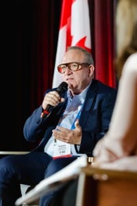 A man in glasses speaking into a microphone during an event photography panel discussion with the Canadian flag in the background.