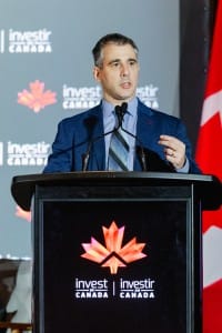 An event photography scene of a man in a suit speaking at a podium with the Invest Canada logo and Canadian flag in the background.