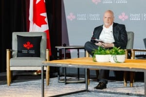 A man sitting onstage with a notepad, beside a chair with the 'invest in Canada' logo, against a backdrop featuring the Canadian flag, captured through event photography.