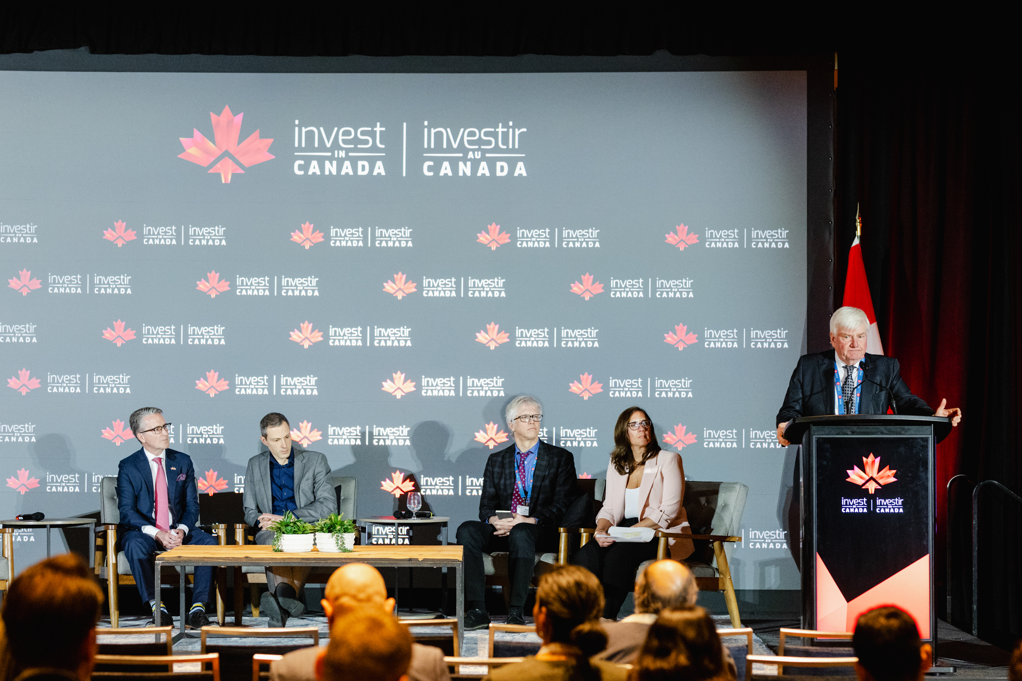 A panel event at Invest Canada with a speaker addressing the audience and four panelists seated onstage, captured by event photography.