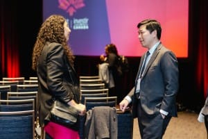 Two professionals conversing in a conference room with empty chairs and a presentation screen in the background, captured during event photography.