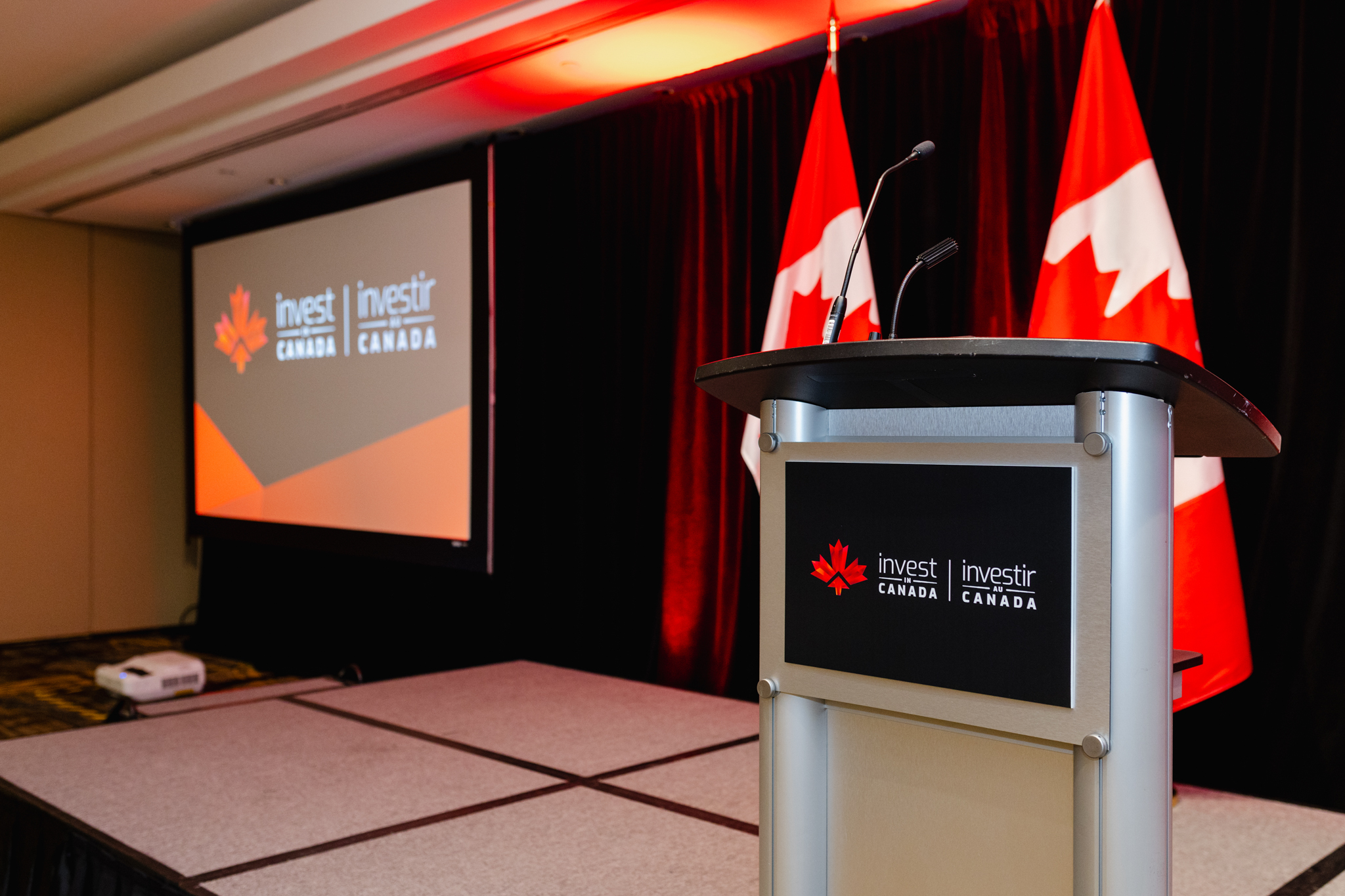 An empty lectern with "Invest in Canada" branding flanked by Canadian flags, featuring event photography, with a projection screen displaying the same logo in the background.