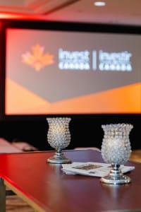 Conference room with decorative table lamps in the foreground and a presentation screen with the words "invest canada" in the background, captured during event photography.