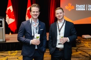 Two men at a conference, holding drinks and wearing id badges, stand in front of banners with Canadian flags, captured in professional event photography.
