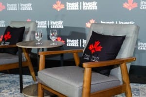 Two empty chairs with cushions, captured in event photography, facing a small table adorned with water glasses, set against a backdrop with the 'invest in canada' logo.