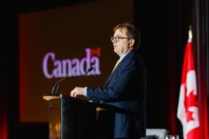 A man in a suit speaking at a podium during an event photography shoot with the word "Canada" projected in the background.