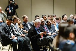 Audience members seated at an event, with one person focused on his smartphone.