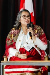 A person sitting on stage speaking into a microphone, wearing a red and gold embroidered shawl over formal attire during event photography.