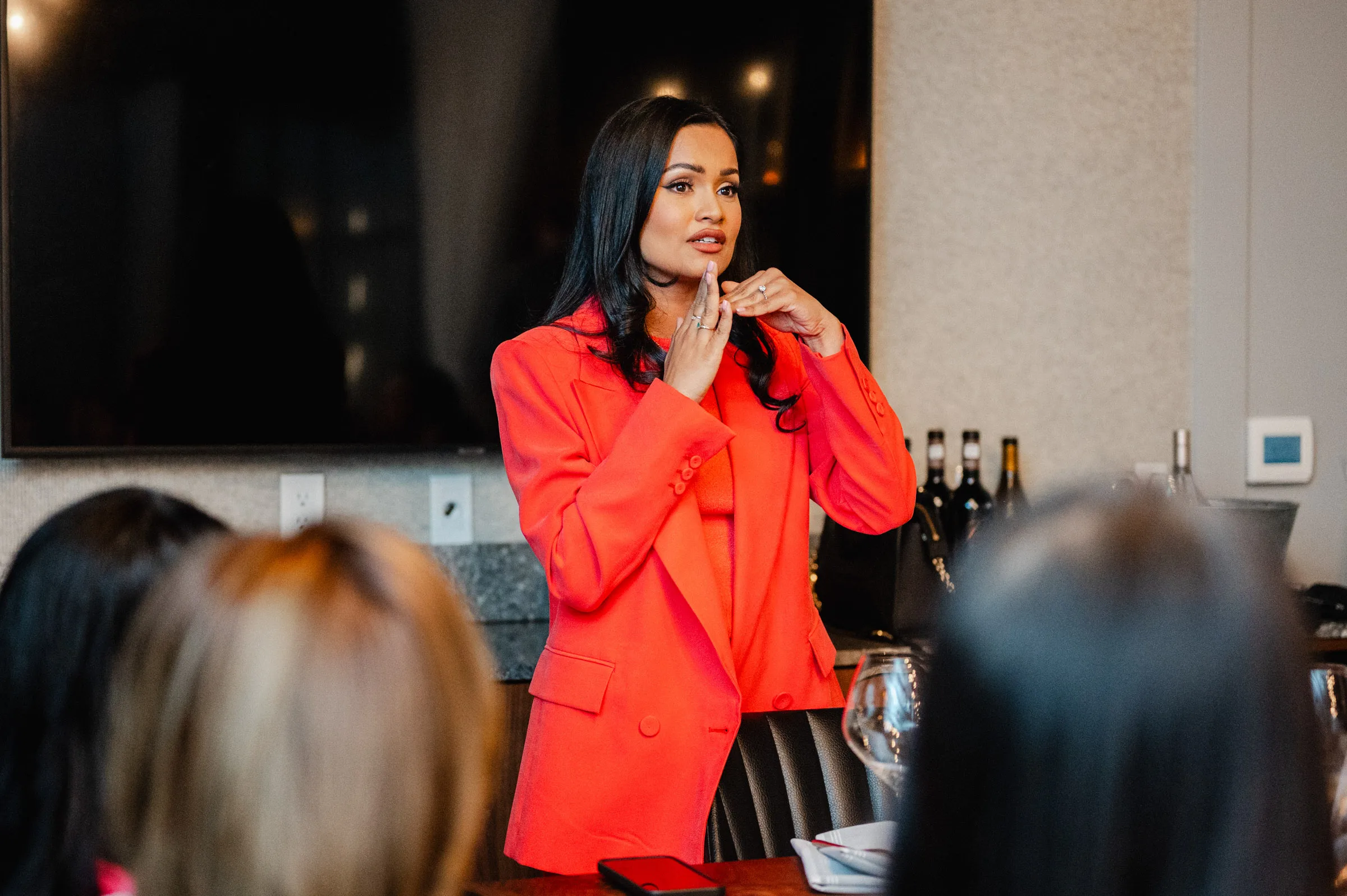 A person in a red blazer addresses an audience during a presentation in a modern indoor setting, perfect for event photography.