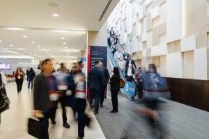 People walking and using an escalator inside a modern, well-lit convention center, with event photography capturing the vibrant signage and displays in the background.