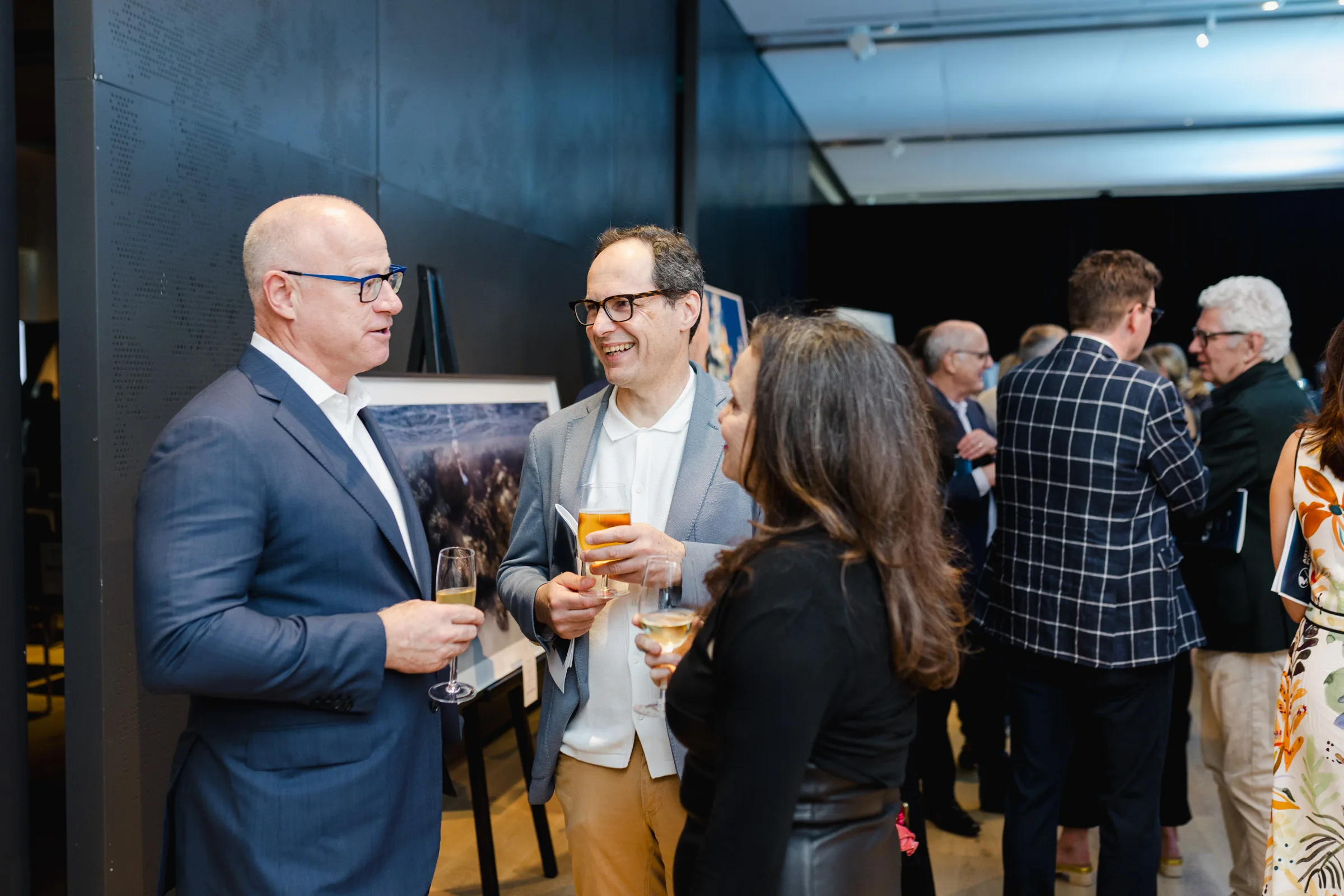 A group of people dressed in business attire stands in a dimly lit room, conversing and holding drinks. Some are focused on conversation, while others are engaged in the background. The scene is captured through expert event photography, highlighting the atmosphere and interactions.