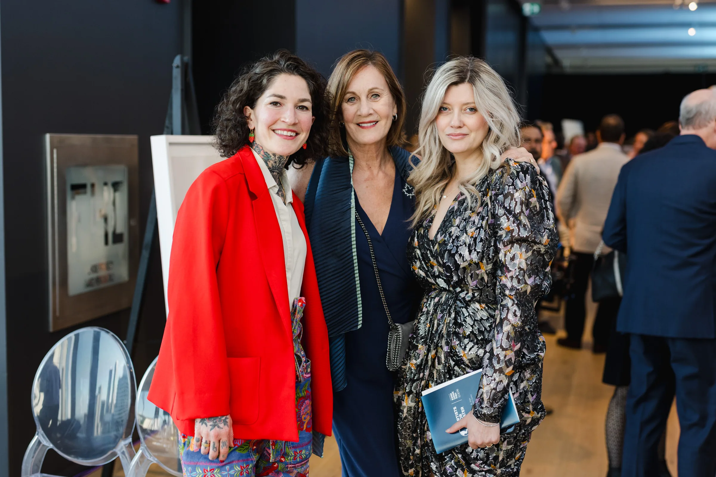 Three women pose together at an event, captured perfectly in event photography; two of them are wearing dresses, while one stands out in a vibrant red blazer.