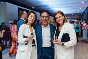 Three people pose and smile for an event photography session at an indoor gathering, with two holding wine glasses. Several other attendees are visible in the background.