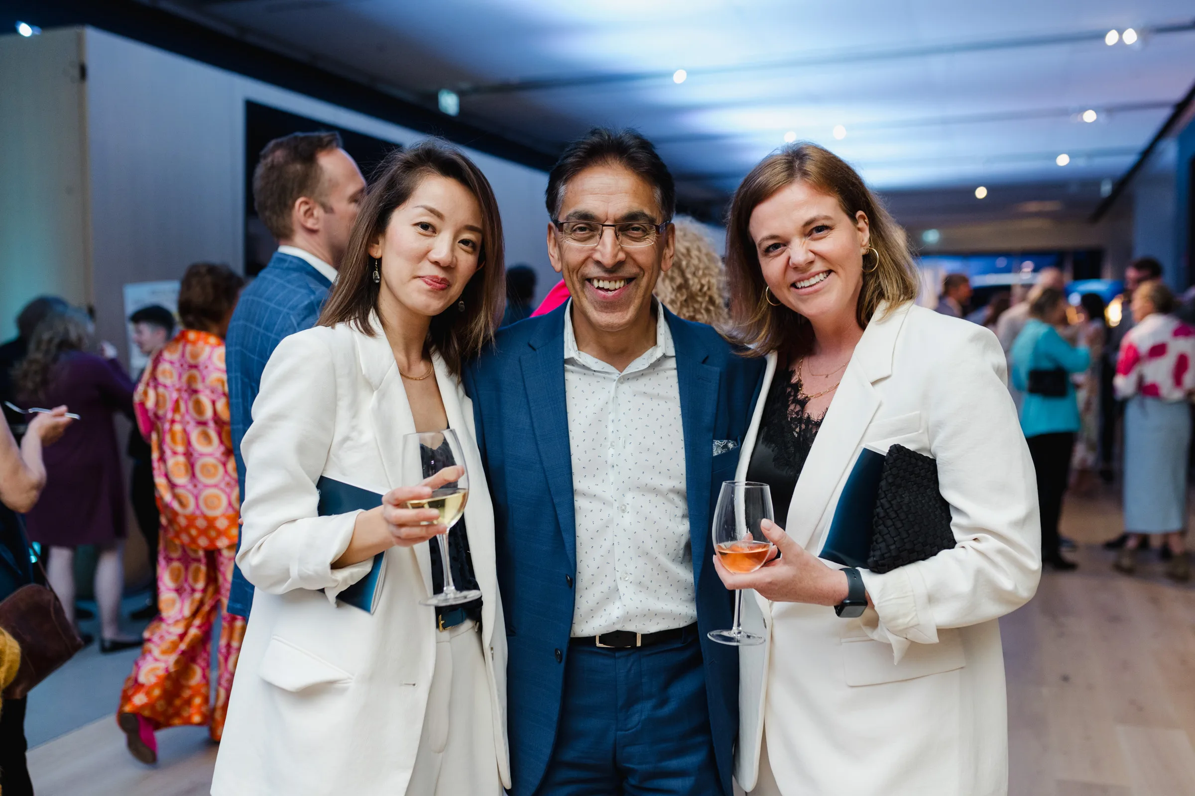 Three people pose and smile for an event photography session at an indoor gathering, with two holding wine glasses. Several other attendees are visible in the background.