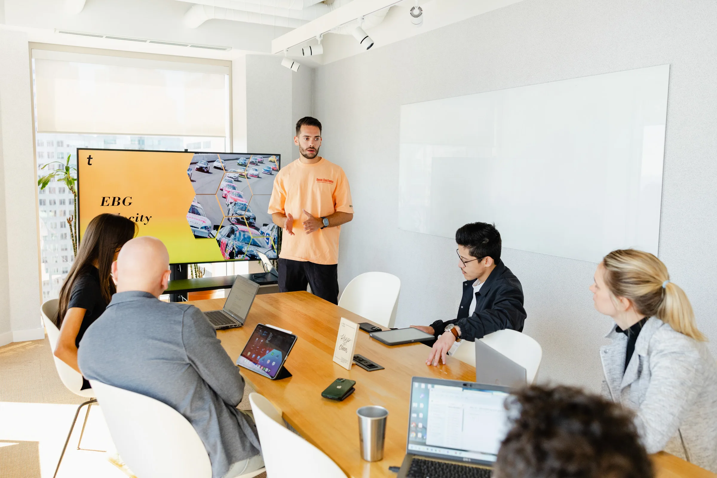 A man stands and presents to four seated colleagues using a TV screen displaying charts and the text “EBG Diversity” in a conference room. Laptops and papers are on the table.