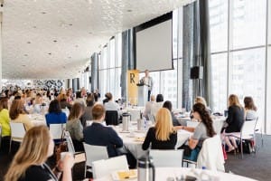 A person is speaking at a podium in front of an audience seated at tables in a large, modern conference room with large windows and city views, perfect for corporate photography.
