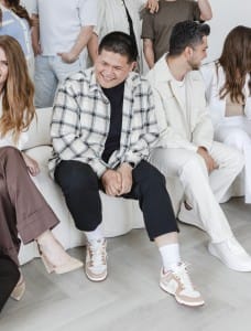 Several people from the creative team are seated indoors, with one person in the center smiling and wearing a plaid shirt. Others on either side are engaged in conversation.