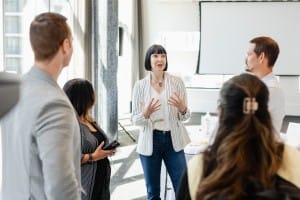 A woman in a striped blazer is speaking to a group of four people indoors, with a projector screen in the background—an ideal scene for corporate photography.