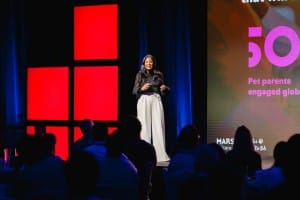 A person stands on a stage presenting to an audience. A large screen beside them displays colorful graphics and text, capturing the essence of corporate photography with its professional visuals.