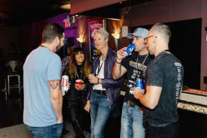 A group of five people standing and holding drinks in a dimly lit room, engaged in conversation near a counter. Perfect for capturing the essence of social event photography.