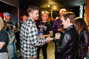 At a vibrant social event, a group mingles indoors, captured through dynamic social event photography. Amidst the lively atmosphere, a man in a plaid shirt hands something to another clad in a leather jacket.