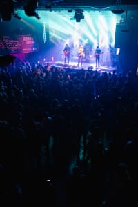 A crowded concert comes alive as the band performs on stage, bathed in blue and purple lights. Capturing the magic of this social event, skilled photographers frame audience members standing and immersed in the electrifying performance.
