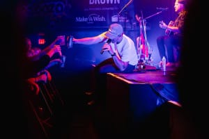 A musician in a cap sits on stage, reaching out with a drink to the audience, captured perfectly by social event photography. In the background, another musician plays guitar, adding to the lively atmosphere.