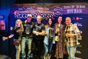 A group of six people, captured by a talented social event photographer, smiles and poses in front of a Curepalooza event banner, holding drinks. They are dressed in casual and event-themed attire.