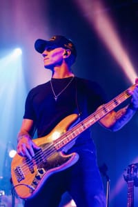 A musician in a cap plays an electric bass guitar on stage with vibrant lighting, capturing the energetic essence of social event photography.