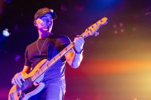 A musician in a black cap and shirt plays a bass guitar on stage under vibrant, colorful lighting, capturing the spirit of social event photography.