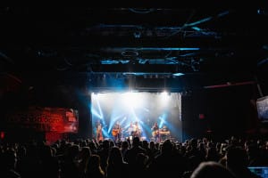A band performs on stage with bright lights, capturing the essence of a social event through vibrant photography as they face a large audience in a dimly lit venue.