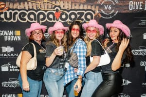Five people wearing pink cowboy hats and glasses pose joyfully in front of a branded event backdrop, embodying the perfect moment of social event photography as they hold drinks and smile at the camera.