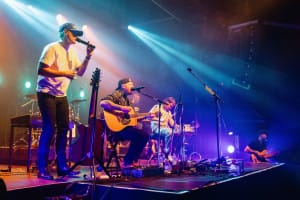 A band performs on stage with various instruments, including guitars and a drum set, captured beautifully in social event photography. Stage lights create a colorful atmosphere.