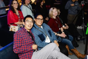 A group of people sits on a couch, smiling and wearing lanyards at an indoor social event, perfectly captured in this vibrant photograph.
