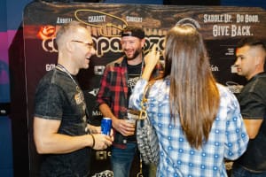 Four people engage in lively conversation in front of a charity concert backdrop. One holds a drink, and another sports a hat, capturing the vibrant essence of social event photography as they enjoy the spirited atmosphere.