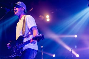 A musician, captured in vibrant Social Event Photography, dons a cap as he sings and plays electric guitar on stage amidst colorful lights.