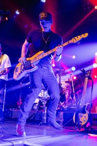 At a vibrant social event, the musician plays an electric bass guitar on stage under purple and red lighting. Wearing a hat and jeans, he stands near a drum set and microphones, captured perfectly in lively photography that highlights the energy of the moment.