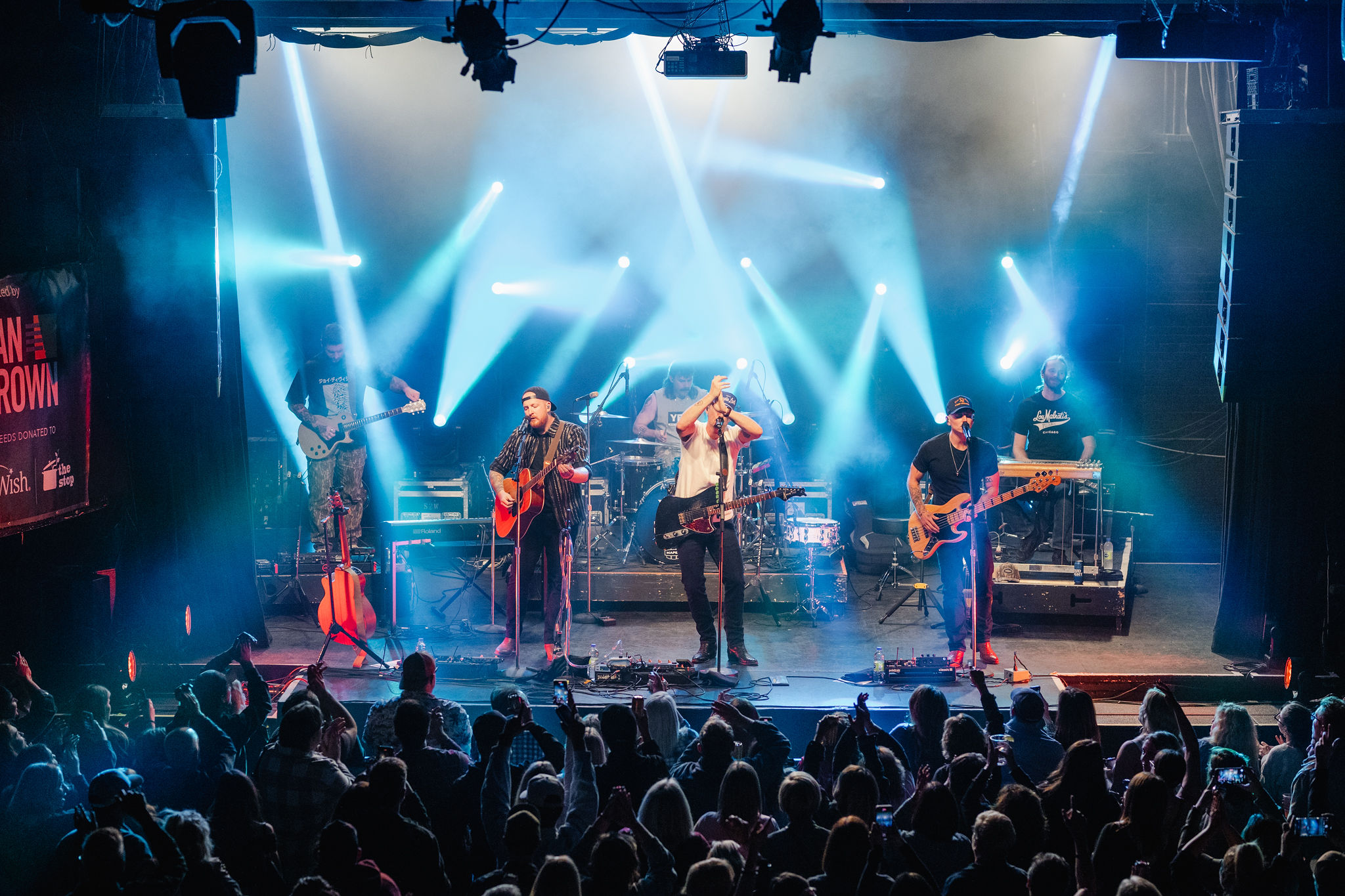 Colorful lights illuminate the stage as a band performs, capturing the essence of a vibrant social event. The cheering audience adds energy while musicians skillfully play guitars, drums, and keyboards. It's a perfect moment for photography that encapsulates the night’s electrifying atmosphere.