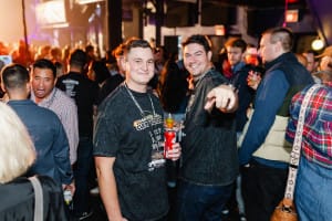 At a bustling social event, two people stand out in the crowd; one playfully points at the camera while holding a drink, capturing the essence of social event photography. In the background, guests continue to mingle and enjoy each other's company.