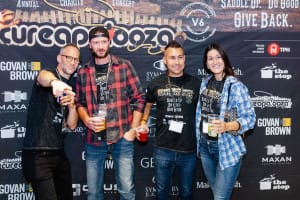 Four people stand in front of a Cureapalooza backdrop, holding drinks and smiling for the camera. They're dressed in casual outfits, featuring plaid shirts and event t-shirts—a perfect snapshot of social event photography capturing joyous memories.