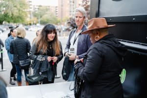 People standing at an outdoor table, captured through the lens of social event photography, engage with a vendor. The urban backdrop, adorned with trees and buildings, frames this lively scene.