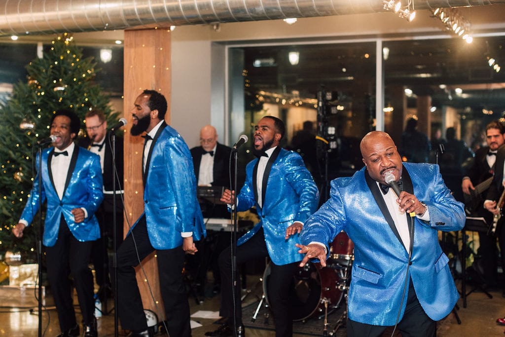 A band performs on stage, with four singers in blue jackets and black pants near a decorated Christmas tree, and musicians playing instruments in the background.