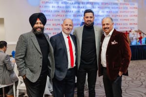 Four men stand together in a group, smiling at an indoor event with a RE/MAX background.