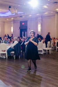 A person in a black dress walks across a room with seated people at tables in the background.