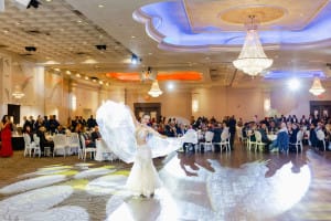 A dancer performs in a large, elegant ballroom with a full audience seated at round tables. Bright lights illuminate the scene.